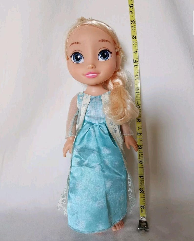 Op de afbeelding: Een blonde pop die een lichtblauwe jurk draagt met een sneeuwvlokkenpatroon. De pop heeft blauwe ogen en een roze mond. De pop is ongeveer 30 cm lang.