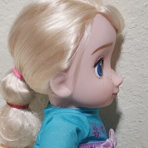 Puede incluir: Perfil de una mu&ntilde;eca con cabello rubio recogido en una cola de caballo. La mu&ntilde;eca tiene ojos azules, pecas y viste un vestido turquesa con un lazo morado. El cabello est&aacute; atado con bandas rosas y rojas.