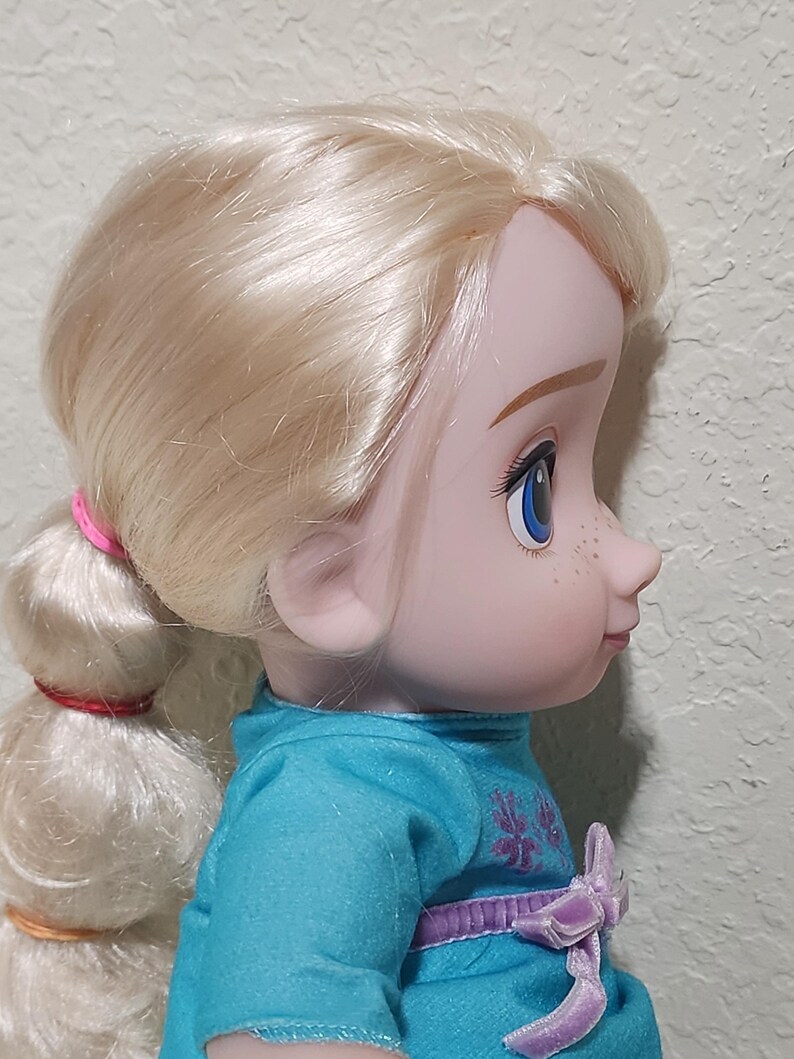 Puede incluir: Perfil de una mu&ntilde;eca con cabello rubio recogido en una cola de caballo. La mu&ntilde;eca tiene ojos azules, pecas y viste un vestido turquesa con un lazo morado. El cabello est&aacute; atado con bandas rosas y rojas.