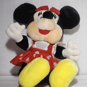 May include: A plush Minnie Mouse doll with black ears, a red bow, and a red and white polka dot dress. The doll has white gloves and yellow feet, with a smiling expression and one hand raised in a thumbs-up gesture.