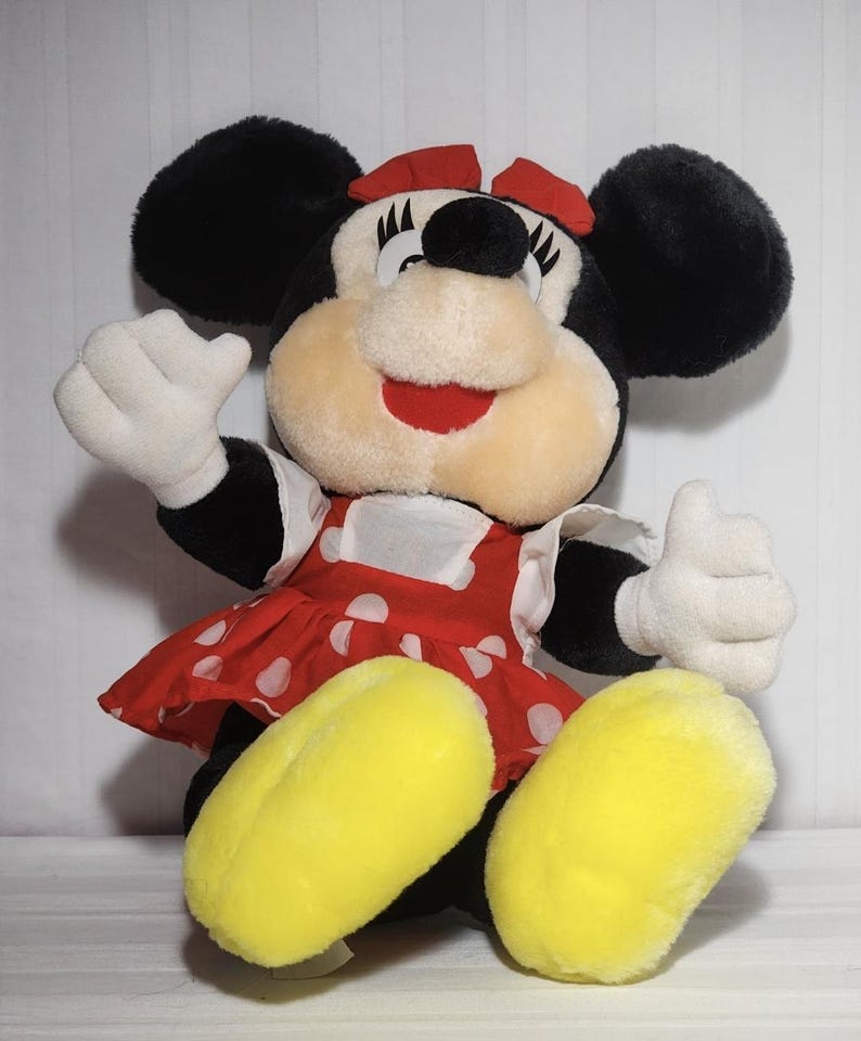 May include: A plush Minnie Mouse doll with black ears, a red bow, and a red and white polka dot dress. The doll has white gloves and yellow feet, with a smiling expression and one hand raised in a thumbs-up gesture.