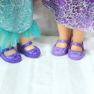 May include: Two pairs of purple doll shoes with bow accents. The shoes have a Mary Jane style with a strap across the top. The dolls are wearing dresses with teal and purple fabric.
