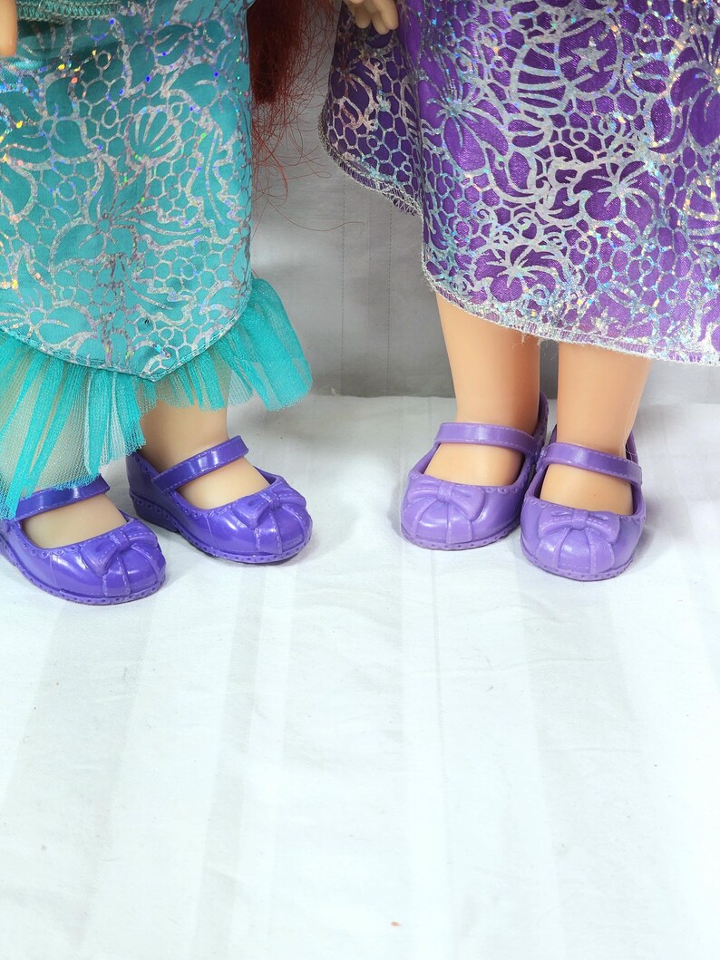 May include: Two pairs of purple doll shoes with bow accents. The shoes have a Mary Jane style with a strap across the top. The dolls are wearing dresses with teal and purple fabric.