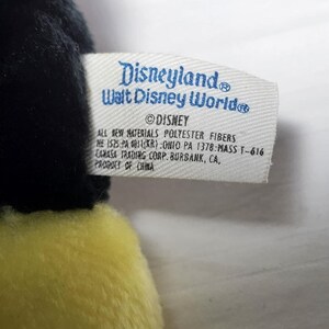 May include: A close-up of a plush toy with a tag that reads "Disneyland Walt Disney World." The tag also includes the Disney logo and information about the materials and manufacturer. The toy has black and yellow fur.
