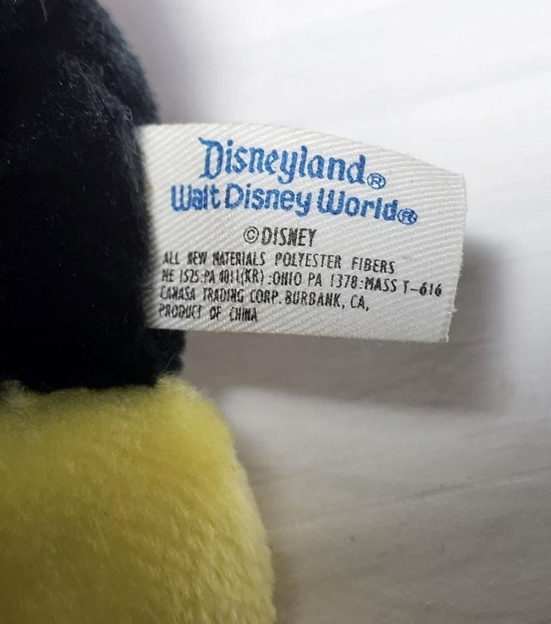 May include: A close-up of a plush toy with a tag that reads "Disneyland Walt Disney World." The tag also includes the Disney logo and information about the materials and manufacturer. The toy has black and yellow fur.