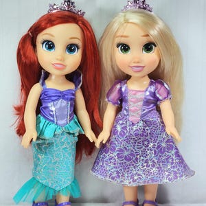 May include: Two dolls of Disney princesses. One doll has red hair, a purple top, and a turquoise mermaid skirt. The other doll has blonde hair and a purple dress. Both dolls wear tiaras and purple shoes. They are holding hands.
