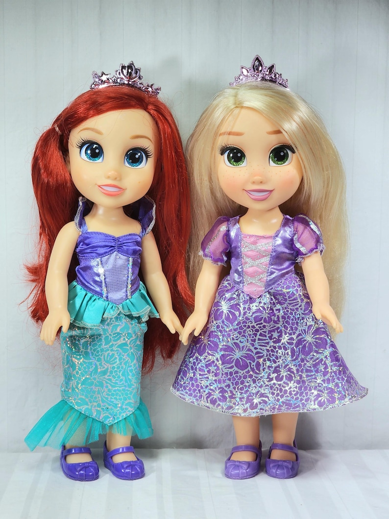 May include: Two dolls of Disney princesses. One doll has red hair, a purple top, and a turquoise mermaid skirt. The other doll has blonde hair and a purple dress. Both dolls wear tiaras and purple shoes. They are holding hands.
