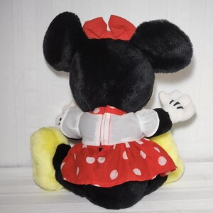May include: A plush Minnie Mouse doll, viewed from the back. The doll has large black ears, a red bow, and a red and white polka-dot dress. White gloves and yellow shoes complete the look. The doll is sitting.
