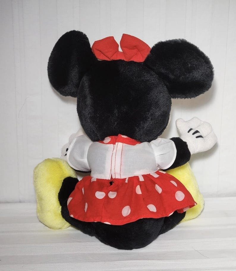 May include: A plush Minnie Mouse doll, viewed from the back. The doll has large black ears, a red bow, and a red and white polka-dot dress. White gloves and yellow shoes complete the look. The doll is sitting.
