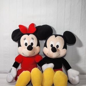 Pu&ograve; includere: Due bambole di peluche dei personaggi Disney, Topolino e Minnie, sedute fianco a fianco. Minnie indossa un vestito e un fiocco rossi, mentre Topolino ha un abito nero. Entrambi hanno grandi orecchie nere, guanti bianchi e scarpe gialle.