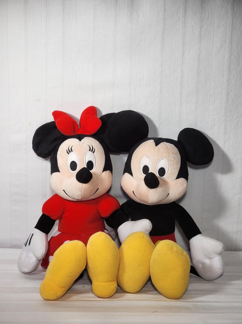 Pu&ograve; includere: Due bambole di peluche dei personaggi Disney, Topolino e Minnie, sedute fianco a fianco. Minnie indossa un vestito e un fiocco rossi, mentre Topolino ha un abito nero. Entrambi hanno grandi orecchie nere, guanti bianchi e scarpe gialle.