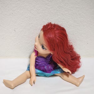 Puede incluir: Mu&ntilde;eca con cabello rojo, vestida con un vestido morado y azul. La mu&ntilde;eca est&aacute; sentada con las piernas extendidas. Tiene ojos azules y piel clara.