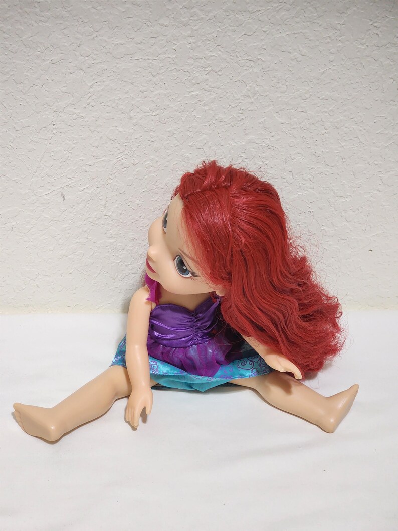 Puede incluir: Mu&ntilde;eca con cabello rojo, vestida con un vestido morado y azul. La mu&ntilde;eca est&aacute; sentada con las piernas extendidas. Tiene ojos azules y piel clara.