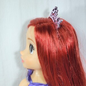 May include: A doll with long, vibrant red hair and a purple dress. The doll has a tiara on its head. The doll's skin is light-toned, and it has large blue eyes and long eyelashes. The doll is a popular character from a children's movie.