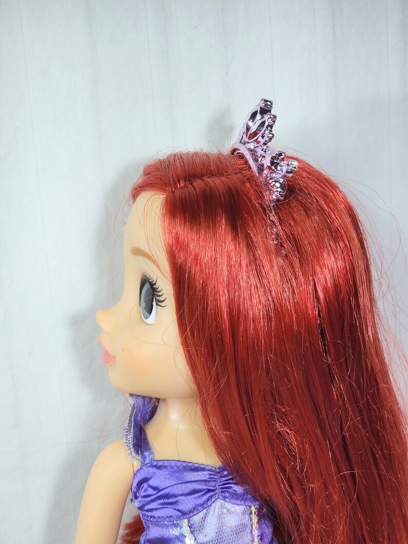 May include: A doll with long, vibrant red hair and a purple dress. The doll has a tiara on its head. The doll's skin is light-toned, and it has large blue eyes and long eyelashes. The doll is a popular character from a children's movie.