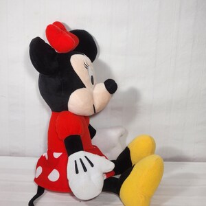 Pu&ograve; includere: Una bambola di peluche di Minnie Mouse seduta. La bambola ha una testa nera con un fiocco rosso, un vestito rosso a pois bianchi, guanti bianchi e scarpe gialle. Un classico personaggio Disney.