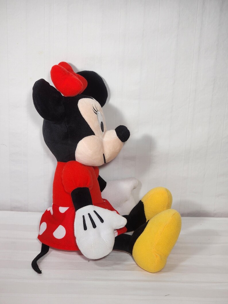 Pu&ograve; includere: Una bambola di peluche di Minnie Mouse seduta. La bambola ha una testa nera con un fiocco rosso, un vestito rosso a pois bianchi, guanti bianchi e scarpe gialle. Un classico personaggio Disney.