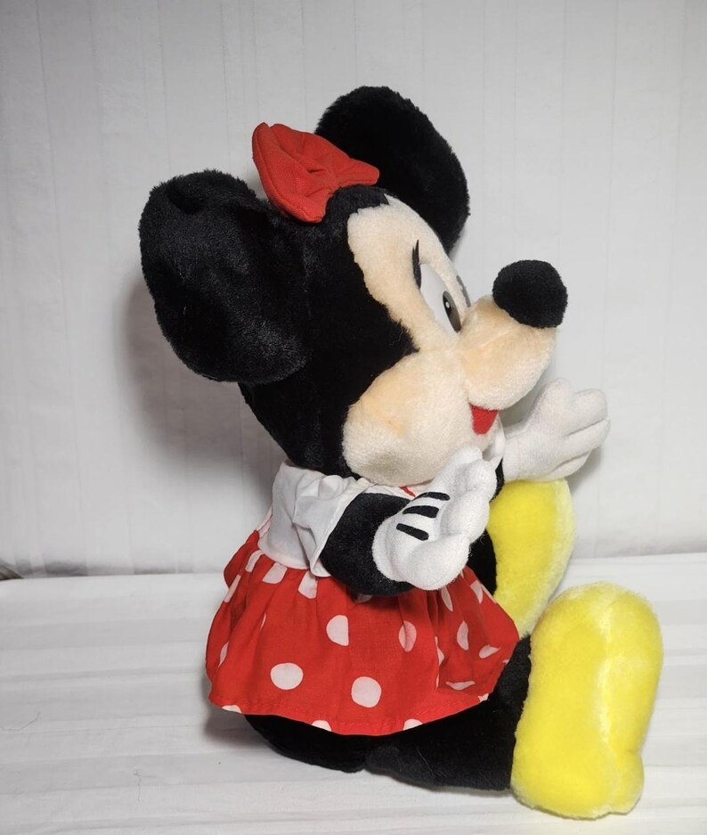 May include: A plush Minnie Mouse doll, featuring black fur, a red polka-dot dress, and a matching bow. The doll has white gloves and yellow shoes. The classic character is in a seated position, with a friendly expression.