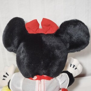 May include: A plush Minnie Mouse doll, viewed from the back, showcasing large black ears and a red bow. The doll wears a white dress with red polka dots and white gloves. The overall design is a classic representation of the iconic character.