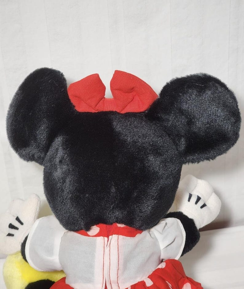 May include: A plush Minnie Mouse doll, viewed from the back, showcasing large black ears and a red bow. The doll wears a white dress with red polka dots and white gloves. The overall design is a classic representation of the iconic character.