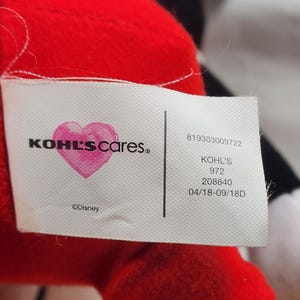 Pu&ograve; includere: Primo piano di un peluche rosso con un'etichetta bianca. L'etichetta presenta il testo "KOHL'S cares." con un cuore rosa e il logo Disney. Sul'etichetta sono visibili anche testo e numeri aggiuntivi.