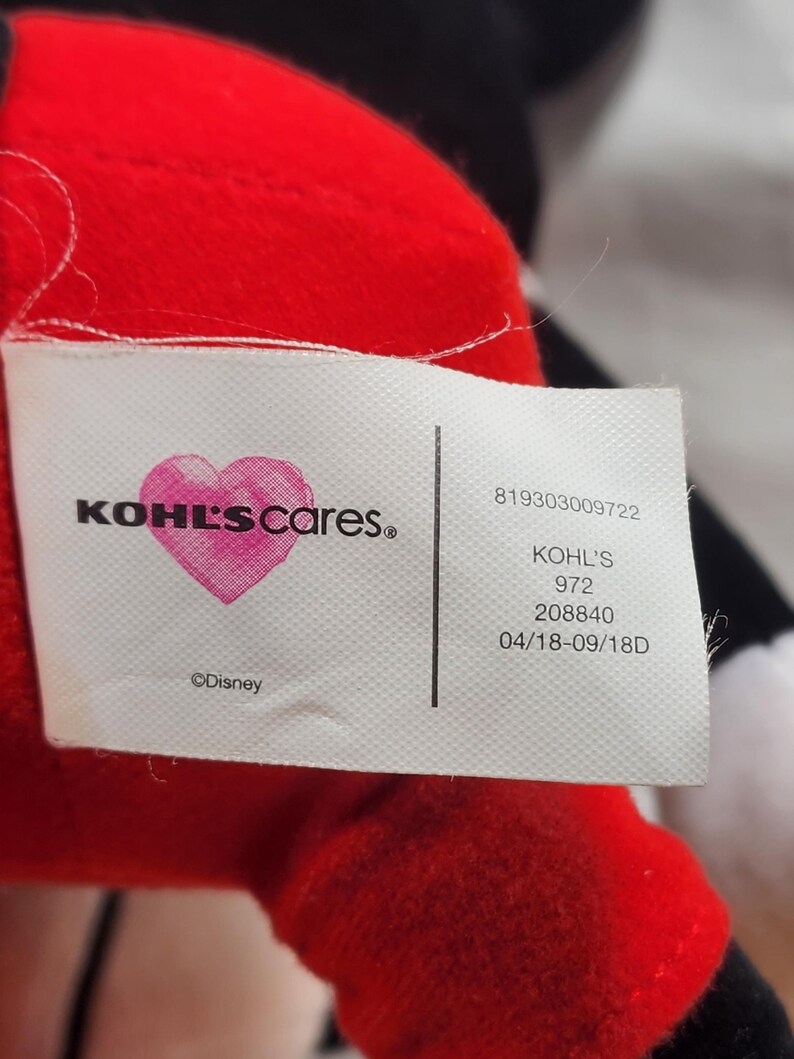 Pu&ograve; includere: Primo piano di un peluche rosso con un'etichetta bianca. L'etichetta presenta il testo "KOHL'S cares." con un cuore rosa e il logo Disney. Sul'etichetta sono visibili anche testo e numeri aggiuntivi.