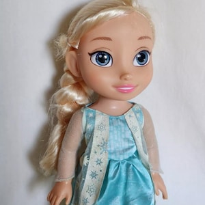 Op de afbeelding: Een blonde pop die een blauwe jurk draagt met een wit sneeuwvlokkenpatroon en een transparante witte overlay. De pop heeft blauwe ogen en een roze glimlach.