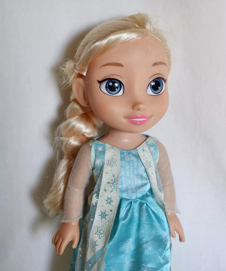 Op de afbeelding: Een blonde pop die een blauwe jurk draagt met een wit sneeuwvlokkenpatroon en een transparante witte overlay. De pop heeft blauwe ogen en een roze glimlach.