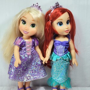 May include: Two Disney princess dolls, Rapunzel and Ariel, stand side-by-side. Rapunzel has long blonde hair, a purple dress, and a tiara. Ariel has red hair, a teal and purple dress, and a tiara. Both dolls wear purple shoes.