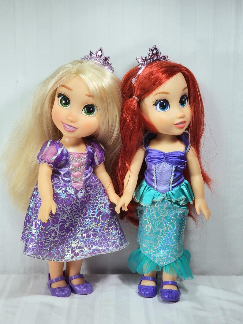 May include: Two Disney princess dolls, Rapunzel and Ariel, stand side-by-side. Rapunzel has long blonde hair, a purple dress, and a tiara. Ariel has red hair, a teal and purple dress, and a tiara. Both dolls wear purple shoes.
