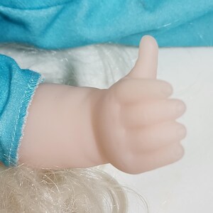 Puede incluir: Una mano de mu&ntilde;eca haciendo un gesto de pulgar hacia arriba. La mano es de color rosa p&aacute;lido y el pulgar est&aacute; levantado. El pelo rubio de la mu&ntilde;eca y una tela turquesa son visibles en el fondo.