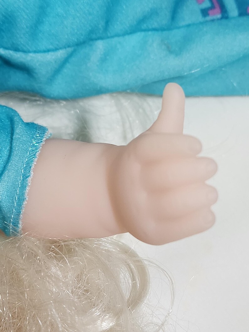 Puede incluir: Una mano de mu&ntilde;eca haciendo un gesto de pulgar hacia arriba. La mano es de color rosa p&aacute;lido y el pulgar est&aacute; levantado. El pelo rubio de la mu&ntilde;eca y una tela turquesa son visibles en el fondo.