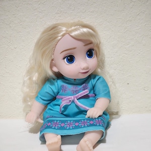 Puede incluir: Mu&ntilde;eca de una ni&ntilde;a con cabello rubio y ojos azules. Viste un vestido turquesa con cintur&oacute;n rosa y ribete floral. La mu&ntilde;eca est&aacute; sentada con las piernas extendidas y los pies visibles.