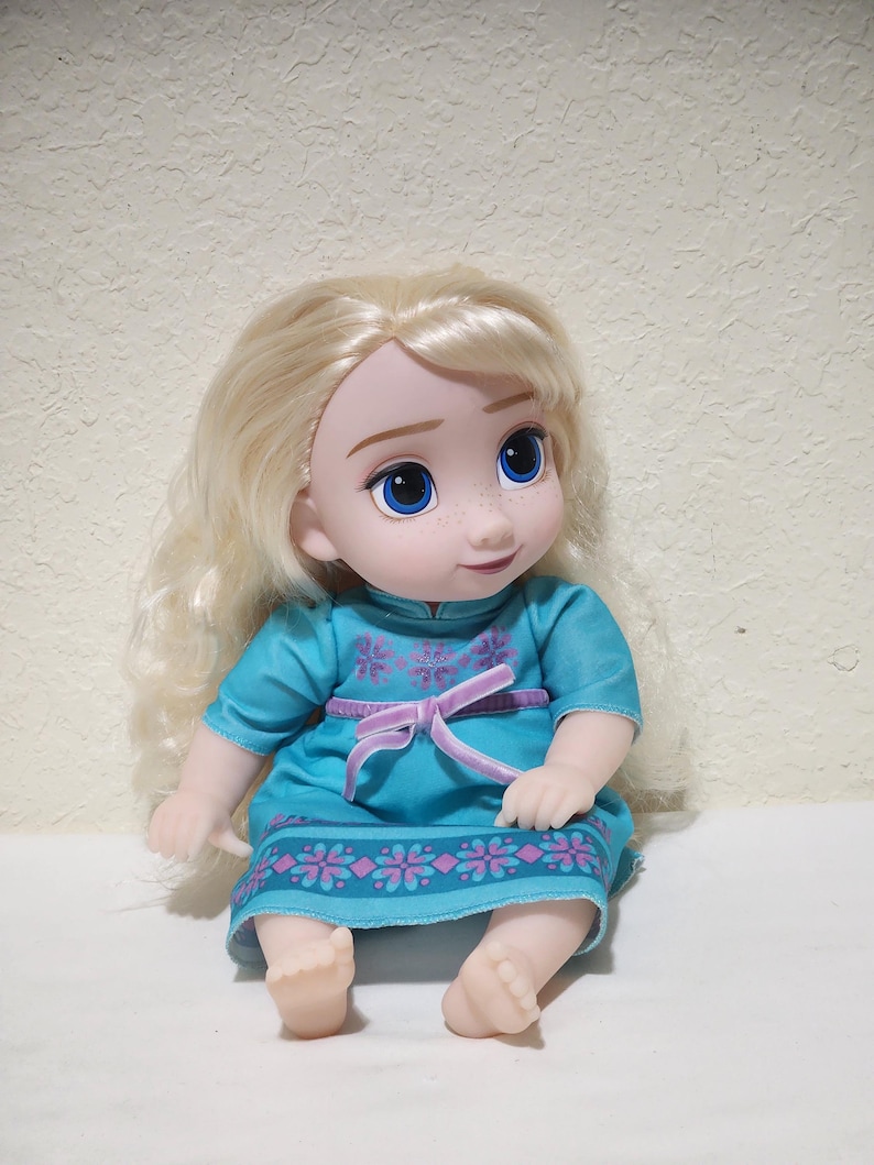 Puede incluir: Mu&ntilde;eca de una ni&ntilde;a con cabello rubio y ojos azules. Viste un vestido turquesa con cintur&oacute;n rosa y ribete floral. La mu&ntilde;eca est&aacute; sentada con las piernas extendidas y los pies visibles.