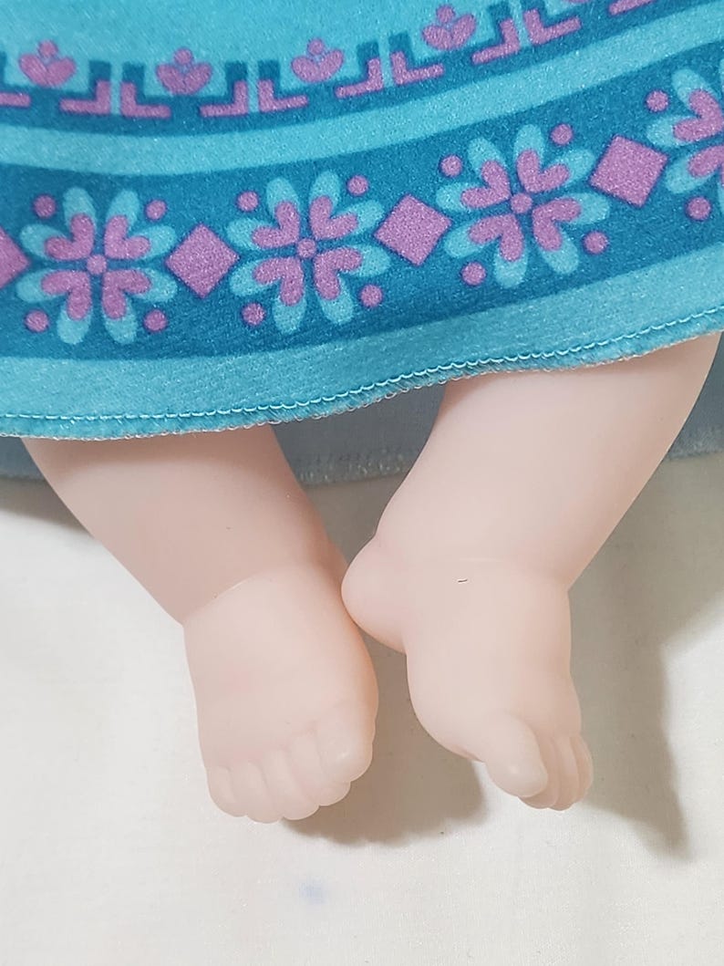 Puede incluir: Primer plano de los pies y las piernas de una mu&ntilde;eca. La piel de la mu&ntilde;eca es de color rosa p&aacute;lido. La mu&ntilde;eca lleva un vestido turquesa con un estampado floral y de diamantes en morado y turquesa.