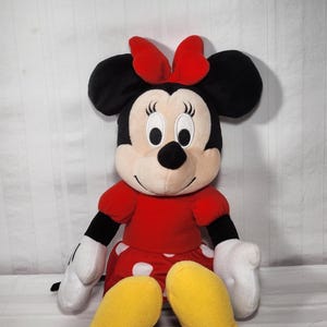 Pu&ograve; includere: Una bambola di peluche di Minnie Mouse, con un vestito rosso a pois bianchi, un fiocco rosso abbinato e grandi orecchie nere. La bambola ha guanti bianchi e scarpe gialle. La bambola &egrave; seduta.