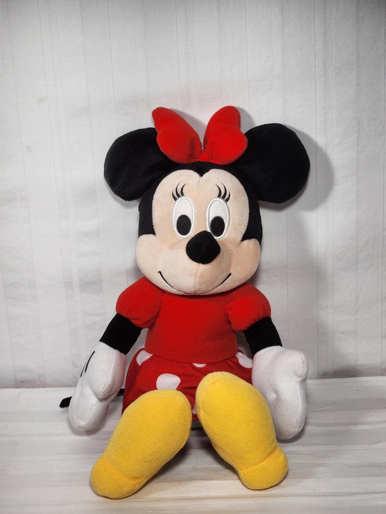 Pu&ograve; includere: Una bambola di peluche di Minnie Mouse, con un vestito rosso a pois bianchi, un fiocco rosso abbinato e grandi orecchie nere. La bambola ha guanti bianchi e scarpe gialle. La bambola &egrave; seduta.