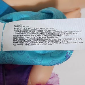 Puede incluir: Primer plano de la ropa de una mu&ntilde;eca y una etiqueta con texto. La etiqueta incluye el logotipo de Disney y varias frases en varios idiomas, incluyendo "Surface Wash" y "Made in China."