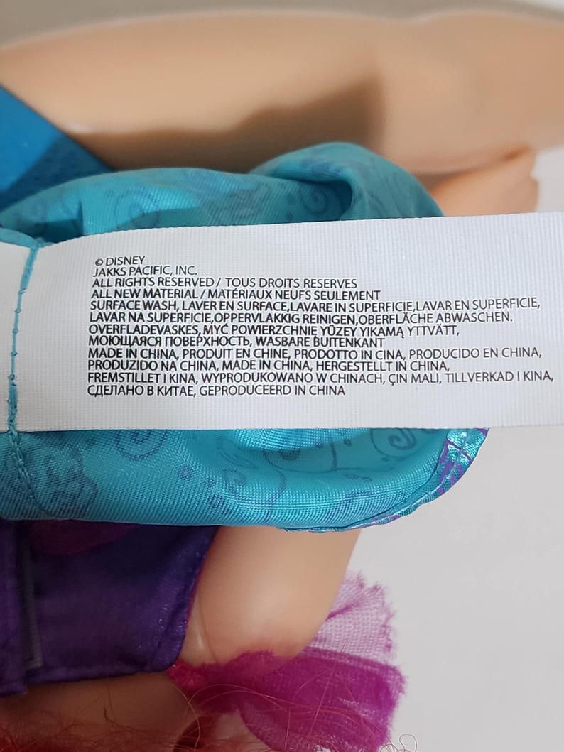 Puede incluir: Primer plano de la ropa de una mu&ntilde;eca y una etiqueta con texto. La etiqueta incluye el logotipo de Disney y varias frases en varios idiomas, incluyendo "Surface Wash" y "Made in China."