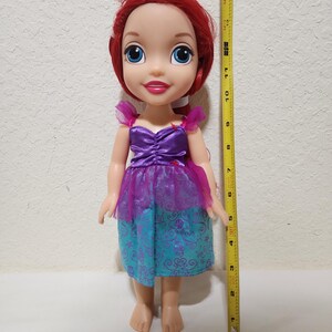 Puede incluir: Mu&ntilde;eca de un personaje con cabello rojo, ojos azules y un vestido morado y turquesa. La mu&ntilde;eca mide aproximadamente 28 cm de altura. El vestido tiene un dise&ntilde;o en capas con un corpi&ntilde;o morado y una falda turquesa.