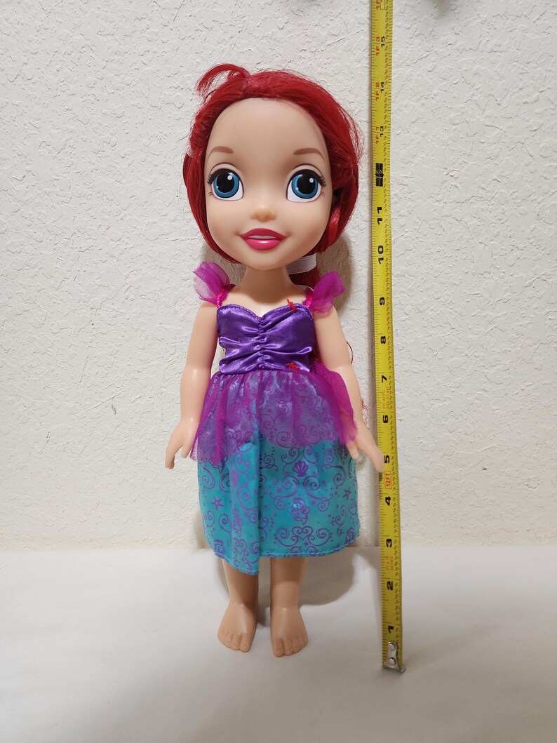 Puede incluir: Mu&ntilde;eca de un personaje con cabello rojo, ojos azules y un vestido morado y turquesa. La mu&ntilde;eca mide aproximadamente 28 cm de altura. El vestido tiene un dise&ntilde;o en capas con un corpi&ntilde;o morado y una falda turquesa.