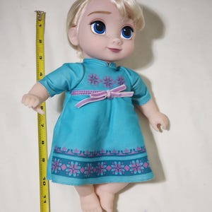 Puede incluir: Mu&ntilde;eca de un personaje con cabello rubio y ojos azules, vestida con un vestido turquesa con estampados florales rosas y un lazo rosa. La mu&ntilde;eca mide aproximadamente 25 cm de alto.