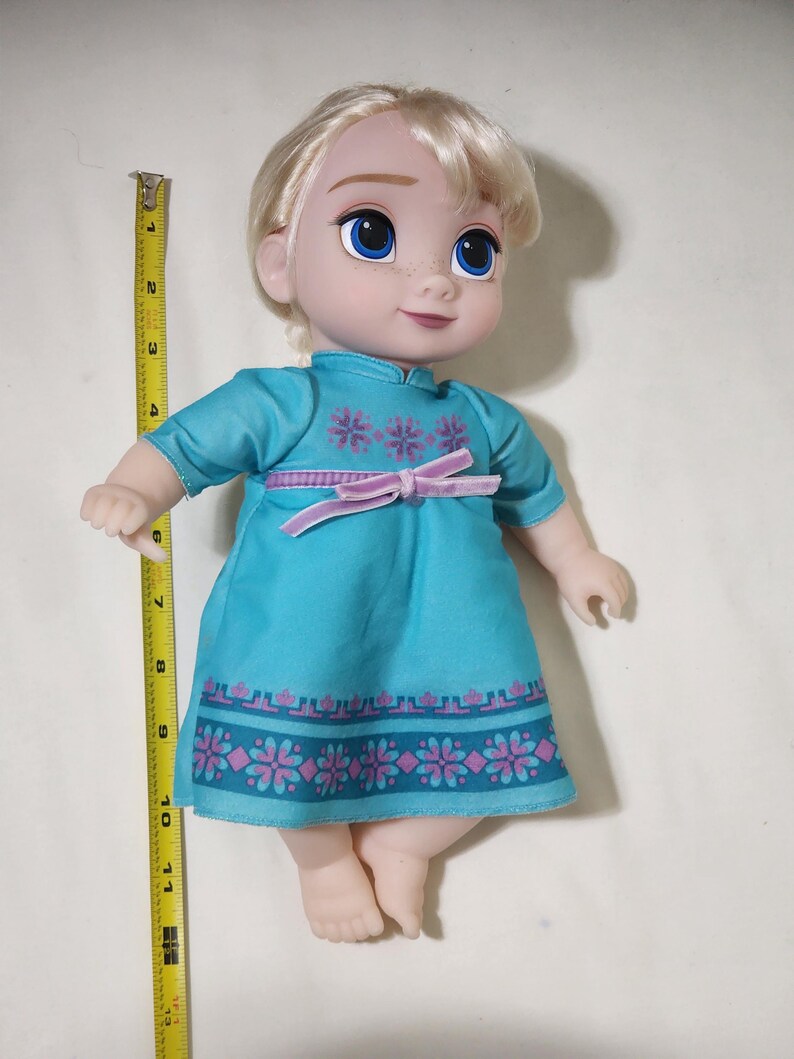 Puede incluir: Mu&ntilde;eca de un personaje con cabello rubio y ojos azules, vestida con un vestido turquesa con estampados florales rosas y un lazo rosa. La mu&ntilde;eca mide aproximadamente 25 cm de alto.