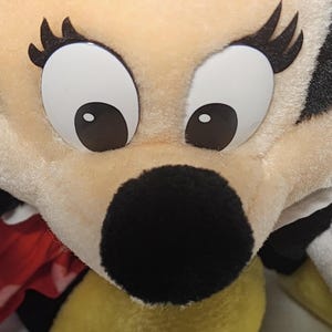 May include: Close-up of a plush toy face with large, white eyes and black pupils. The toy has a black nose and long, black eyelashes. The fur is a light beige color, with a hint of yellow and red fabric visible.