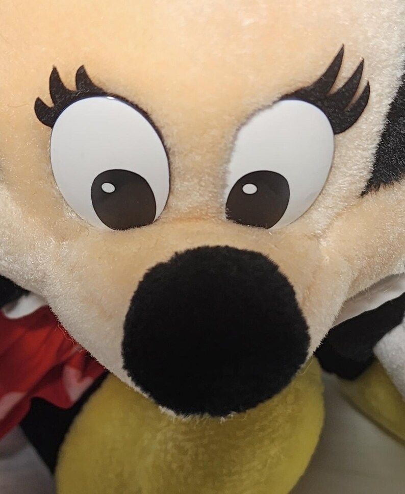 May include: Close-up of a plush toy face with large, white eyes and black pupils. The toy has a black nose and long, black eyelashes. The fur is a light beige color, with a hint of yellow and red fabric visible.