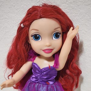 Puede incluir: Mu&ntilde;eca con cabello rojo y ojos azules, vestida con un vestido morado con una superposici&oacute;n de tul rosa. La mu&ntilde;eca tiene una expresi&oacute;n sonriente y un brazo levantado.