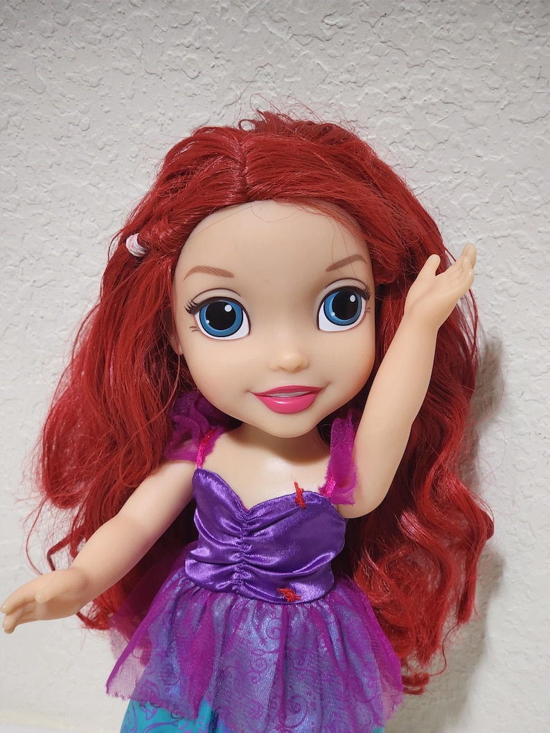 Puede incluir: Mu&ntilde;eca con cabello rojo y ojos azules, vestida con un vestido morado con una superposici&oacute;n de tul rosa. La mu&ntilde;eca tiene una expresi&oacute;n sonriente y un brazo levantado.