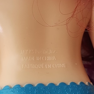 Puede incluir: Primer plano de la espalda de una mu&ntilde;eca, con el texto "JAKKS Pacific, Inc. MADE IN CHINA FABRIQU&Eacute; EN CHINE" impreso en blanco sobre la piel. La mu&ntilde;eca tiene pelo rojo y una falda azul estampada.