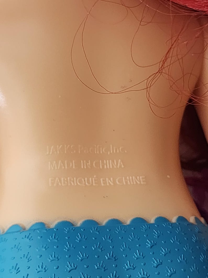 Puede incluir: Primer plano de la espalda de una mu&ntilde;eca, con el texto "JAKKS Pacific, Inc. MADE IN CHINA FABRIQU&Eacute; EN CHINE" impreso en blanco sobre la piel. La mu&ntilde;eca tiene pelo rojo y una falda azul estampada.