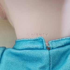 Puede incluir: Primer plano del cuello y los hombros de una mu&ntilde;eca. La mu&ntilde;eca tiene el pelo rubio y lleva un vestido turquesa. La palabra "&copy; DISNEY" est&aacute; impresa en la parte posterior del cuello de la mu&ntilde;eca.